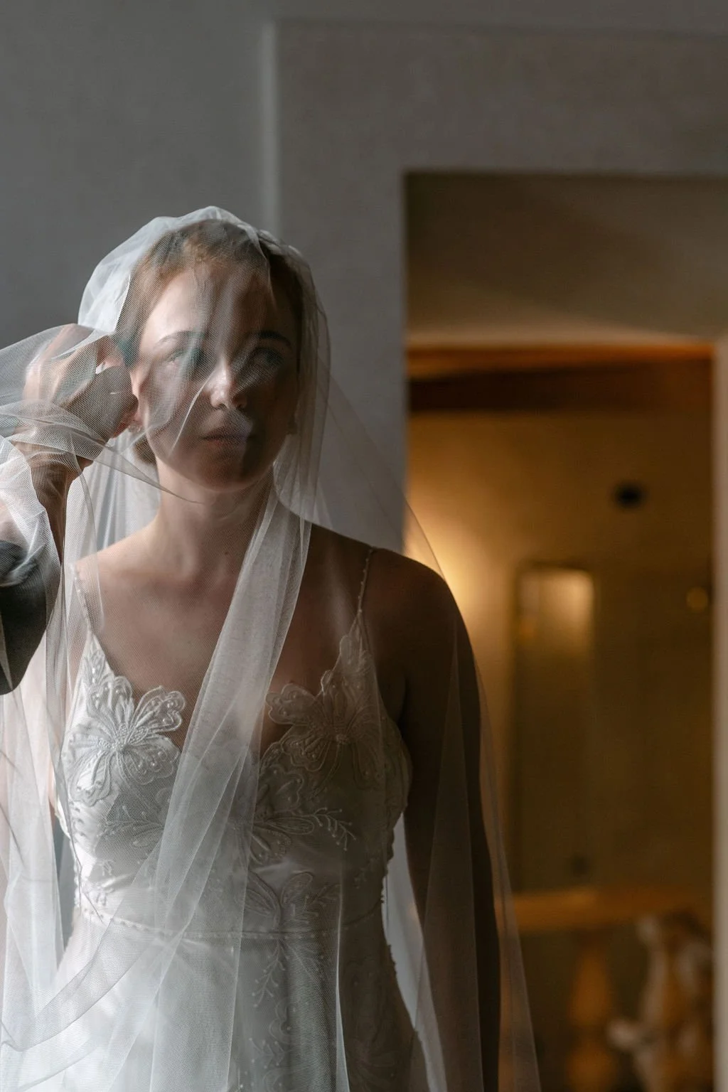 A woman wearing a wedding dress with butterfly embroidery looking into a mirror, partially covered by a veil, with her hand touching her face.
