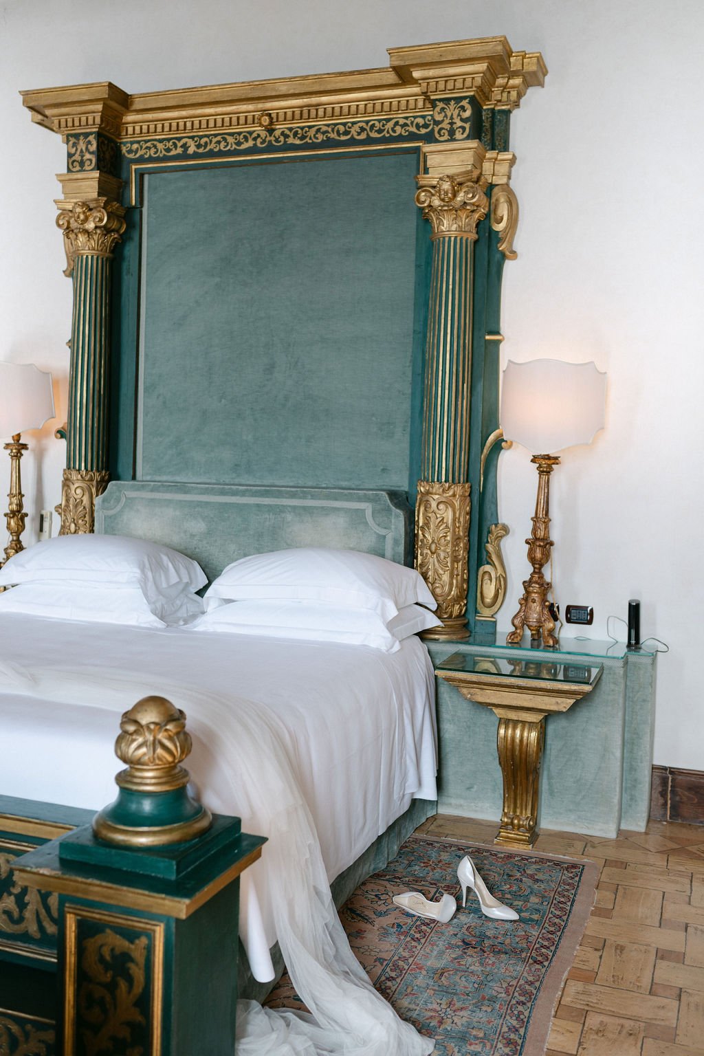 A luxurious bedroom featuring a large bed with white linens and pillows, an ornate headboard, and an elaborately decorated canopy with gold accents. Beside the bed, there are two tall lamps with cream-colored shades, and a small glass-top nightstand 