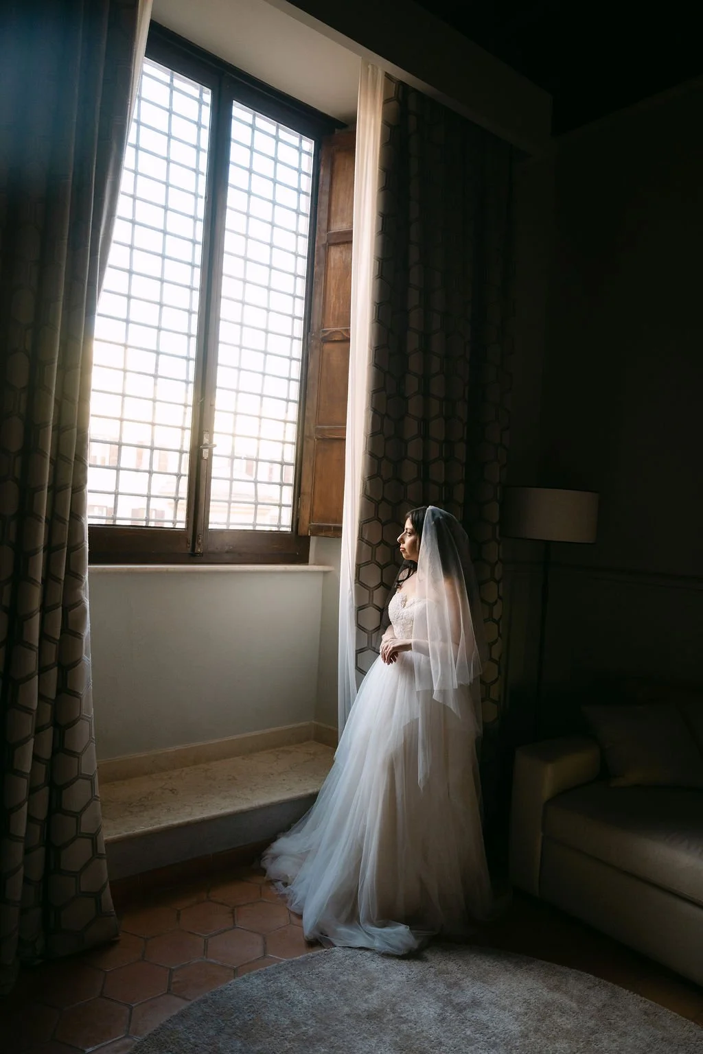 A woman in a wedding dress and veil standing by a large window with curtains, looking outside during daylight in a room with patterned walls and a beige armchair.