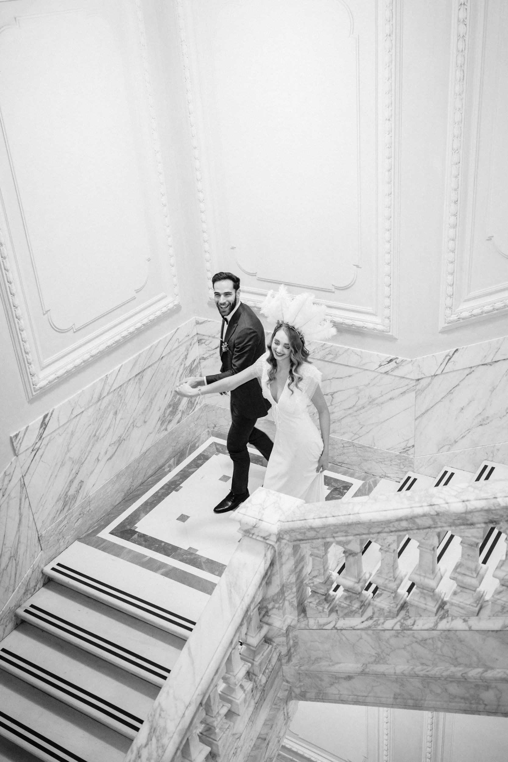 A couple in wedding attire walking up a marble staircase in an elegant, ornate building, smiling and holding hands.