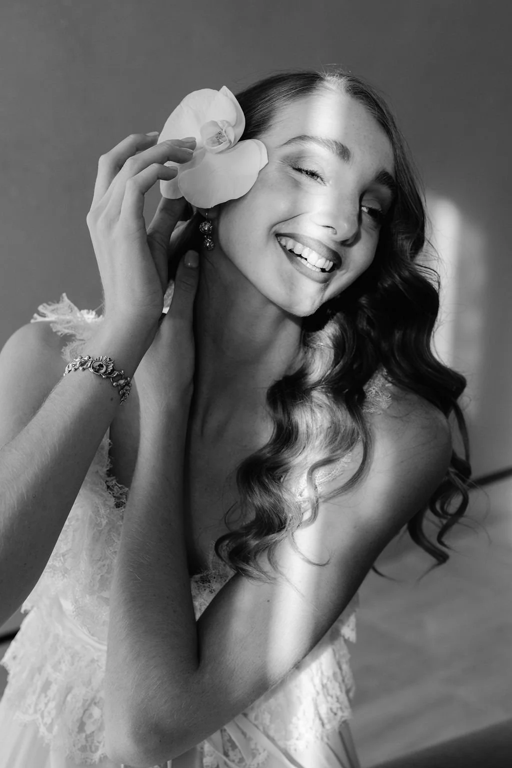 Black-and-white portrait of a young woman with long, wavy hair, smiling with one eye closed, holding a large flower near her face, wearing earrings and a lace top.