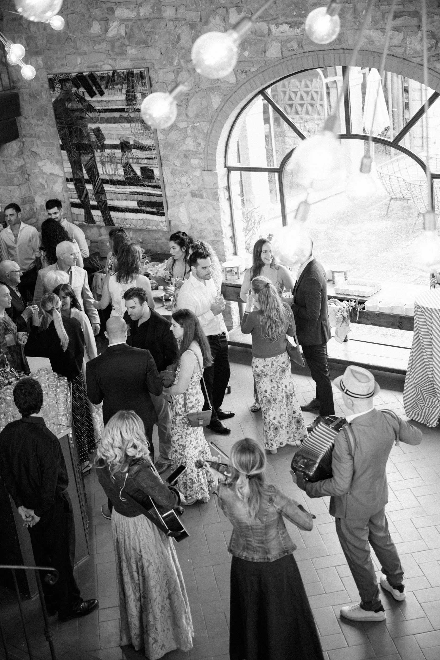 People at a social gathering or party inside a venue with a large window and brick walls, some playing instruments, others talking and mingling.