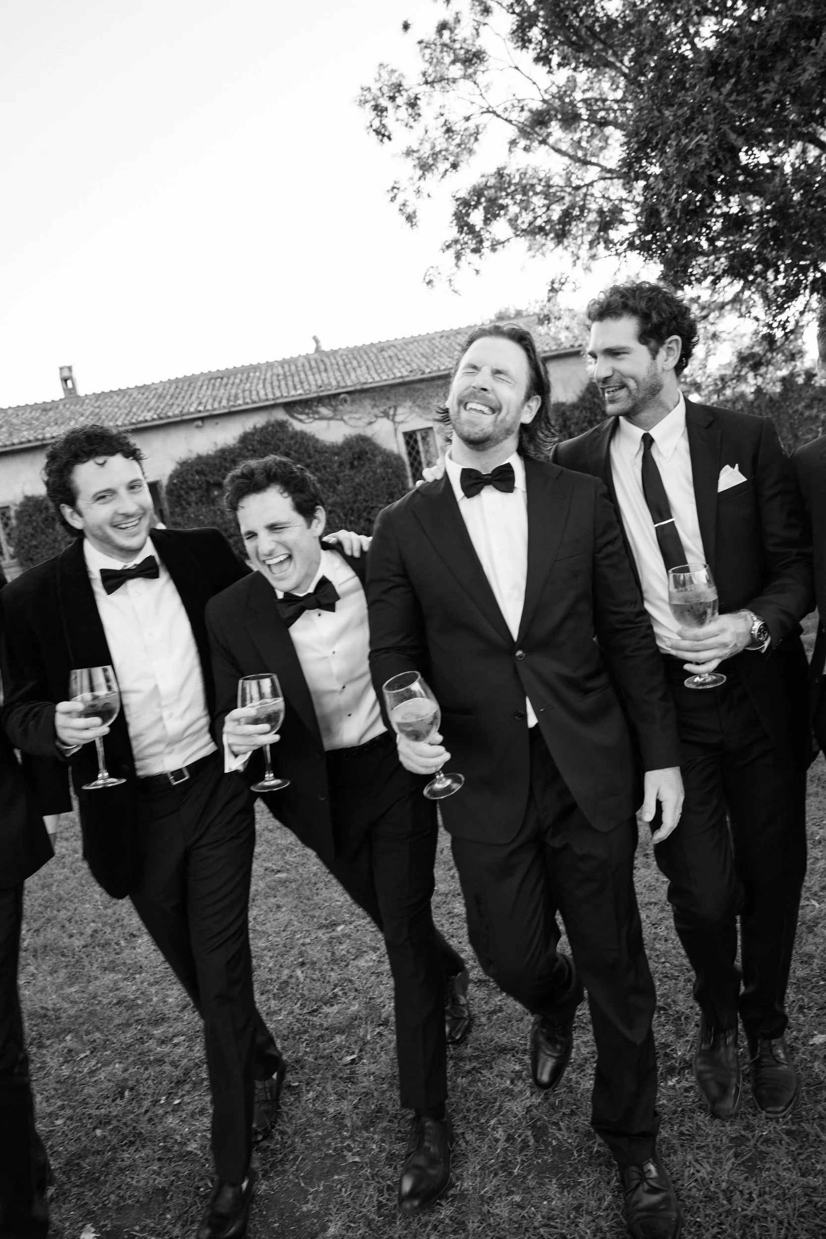 Group of five men in tuxedos and bow ties, holding glasses of wine, smiling and having fun outdoors near a house and trees, black and white photo.
