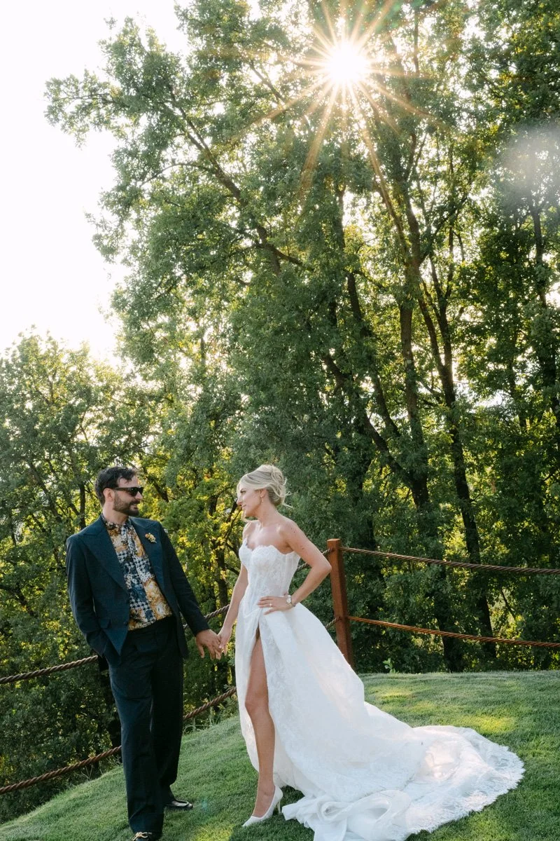 A bride and groom holding hands outdoors on a grassy area, with lush green trees and sunlight shining through the leaves in the background.