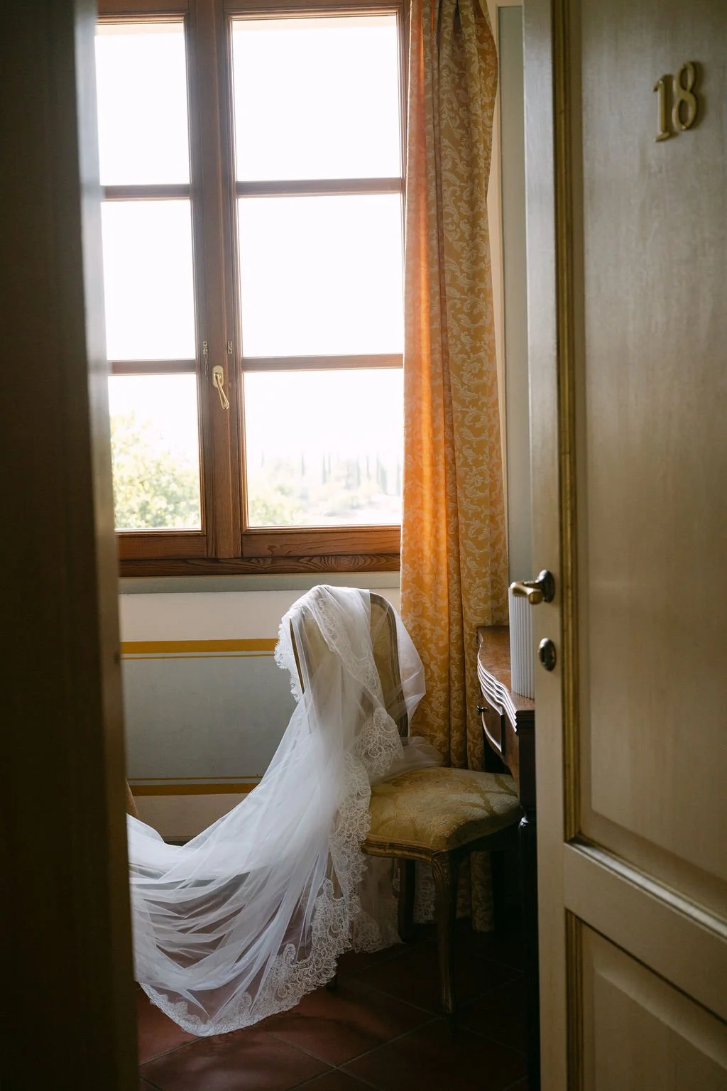 A softly lit room featuring a wooden-framed window with yellow patterned curtains, a vintage chair with a lace cloth draped over it, and the number 18 on the door.