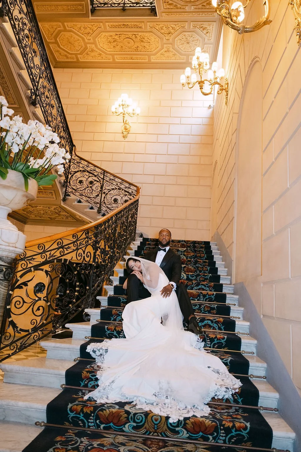 A bride and groom in wedding attire sitting on a grand staircase in an elegant, chandelier-lit interior. The bride is leaning on the groom, who is dressed in a tuxedo, with ornate iron railing and decorative ceiling panels visible.