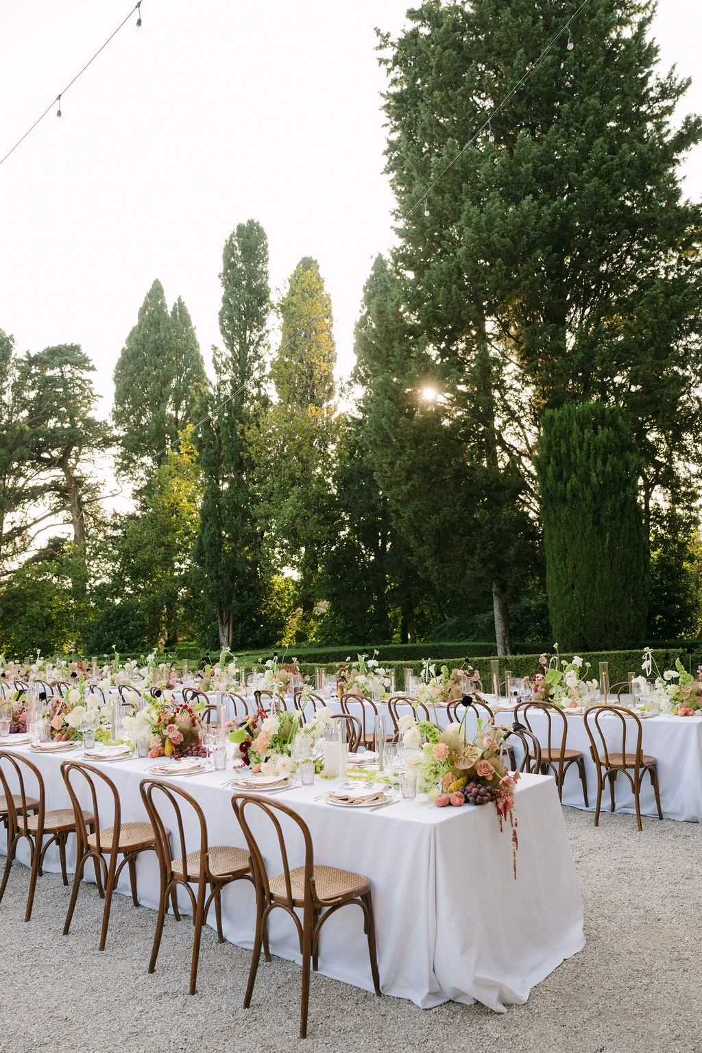 Long outdoor banquet table decorated with floral centerpieces, surrounded by wooden chairs, set in a lush green garden with tall trees and string lights overhead.