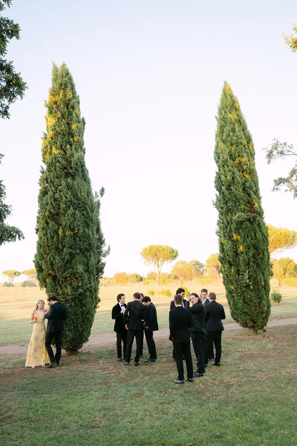 A group of men in tuxedos and women in formal dresses gathered outdoors near tall cypress trees in a park or garden setting.