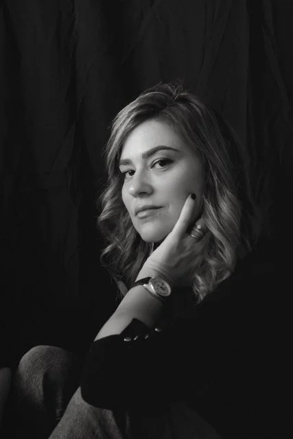 A woman with wavy hair resting her head on her hand, looking at the camera, in black and white.