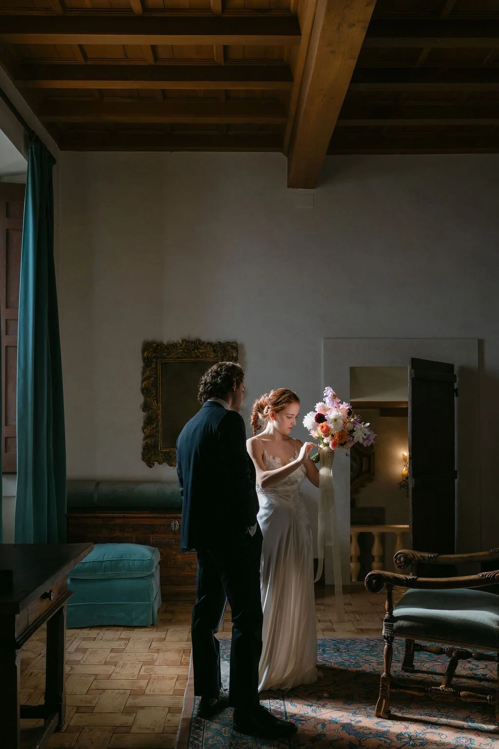 A bride in a white wedding dress holding a bouquet of flowers and a man in a dark suit standing in a room with wooden ceiling beams, vintage furniture, and a decorative mirror.