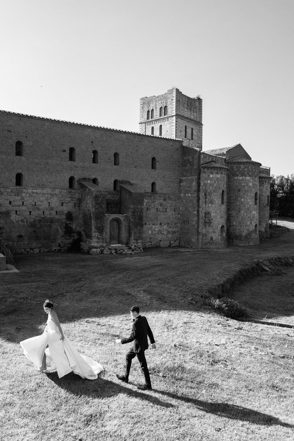 A black-and-white photo of a bride and groom walking on a grassy area in front of a historic castle. The bride wears a flowing white gown, and the groom is dressed in a dark suit. The castle features stone walls and towers, with a large central struc