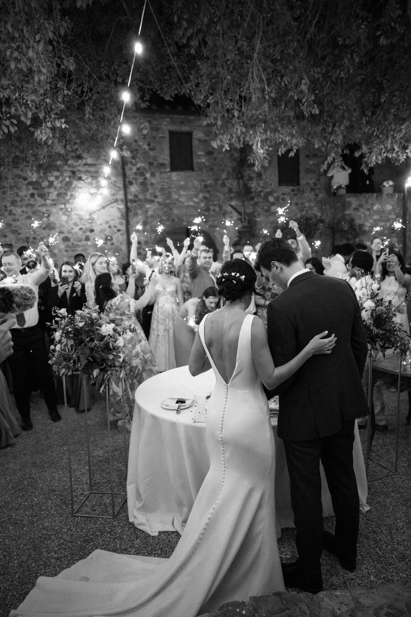 A black and white photo of a wedding reception at night, with a bride and groom standing close together, facing away from the camera. The bride is wearing a white gown with buttons down the back and a floral headpiece, and the groom is in a dark suit