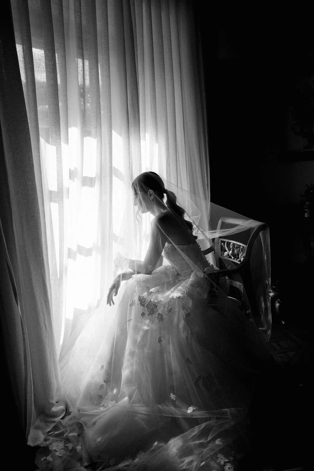 A woman in a wedding dress sits in front of a window with sheer curtains, illuminated by daylight, wearing a veil and wedding jewelry.