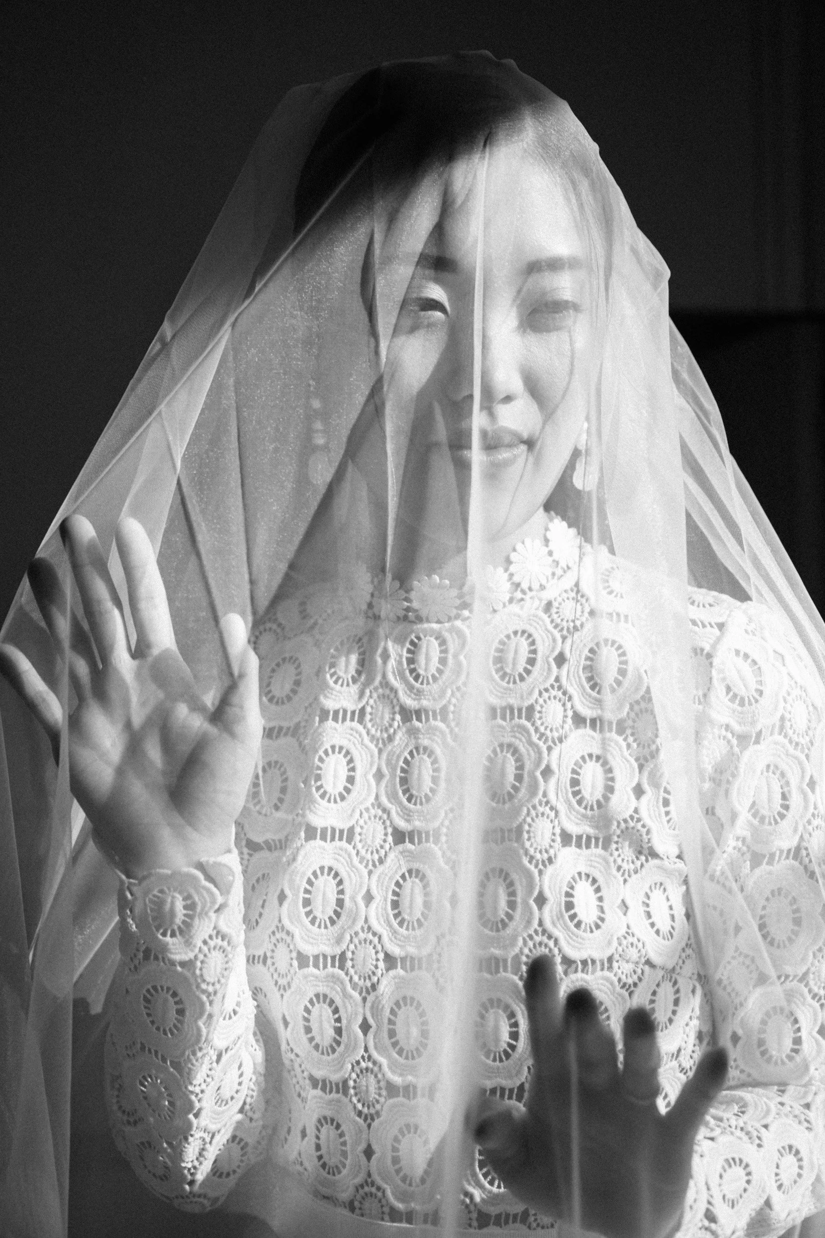 A woman behind a lace veil, smiling and wearing earrings and a patterned dress.
