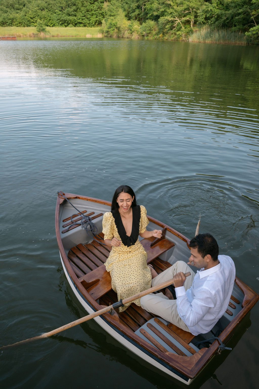 A woman and a man smiling and laughing while rowing a small wooden boat on a calm lake surrounded by greenery.