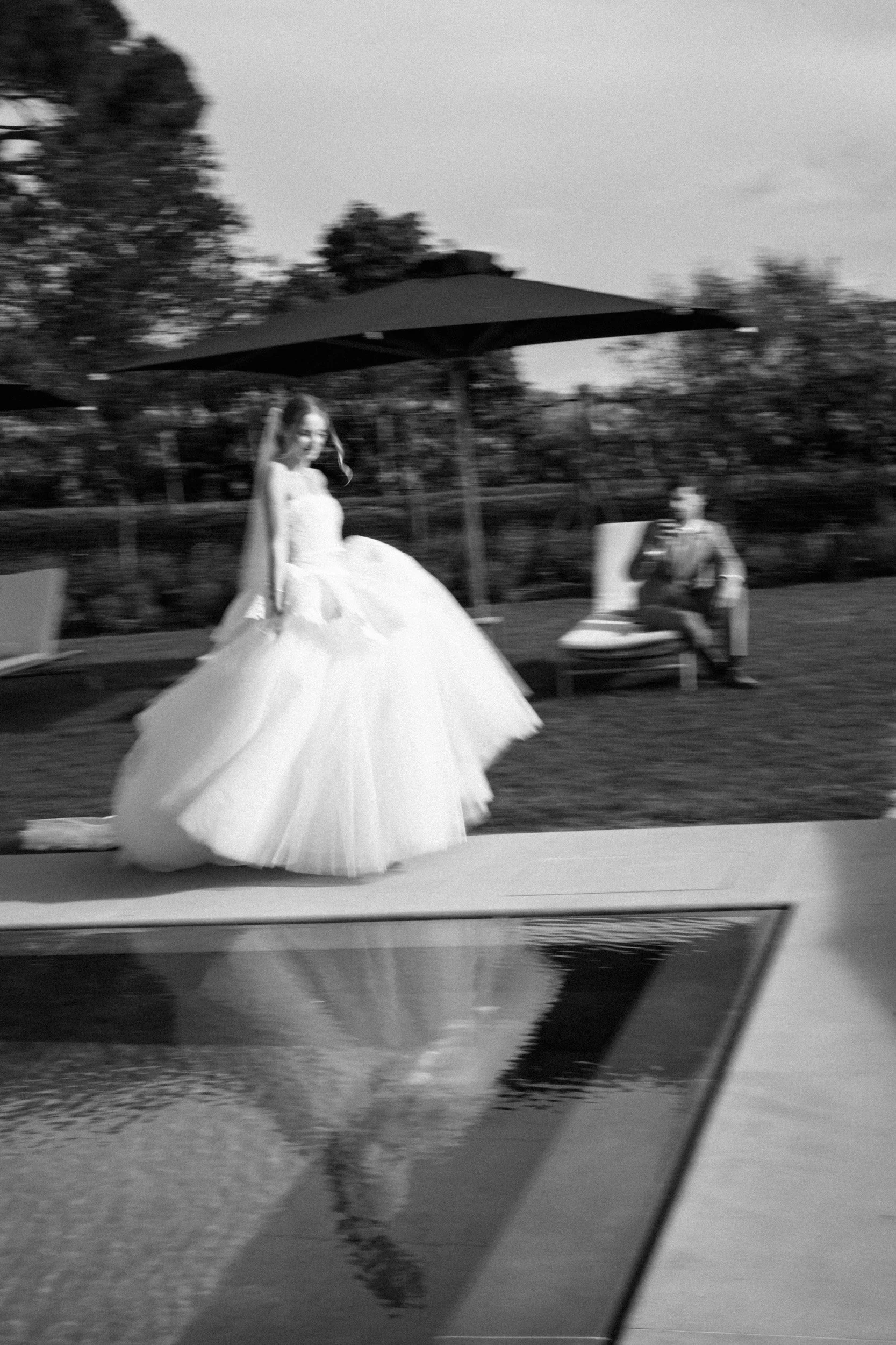 A woman in an elegant white wedding gown standing beside a reflective pool. A man is seated in the background under a large umbrella, possibly looking at his phone.