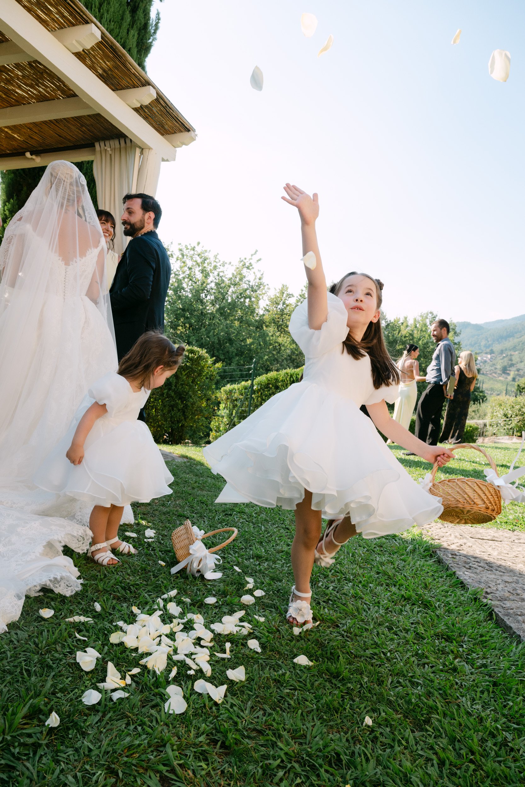 Young girl cheery during a wedding celebration, throwing flower petals while two other children gather petals on the grass, with a couple wedding officiating in the background outdoors.