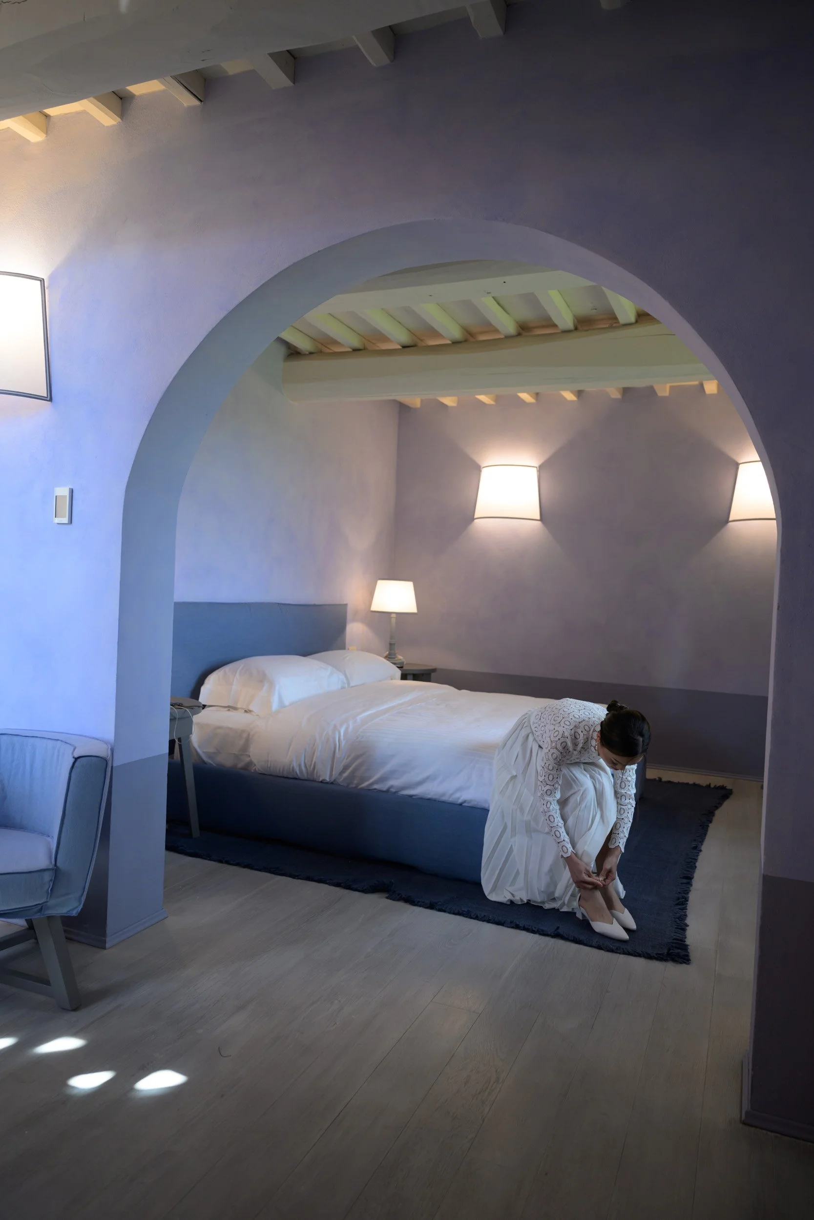 A woman in a white dress is kneeling on a dark rug, tying the laces of her high-heeled shoes, next to a bed in a softly lit bedroom with lavender walls and modern wall sconces.