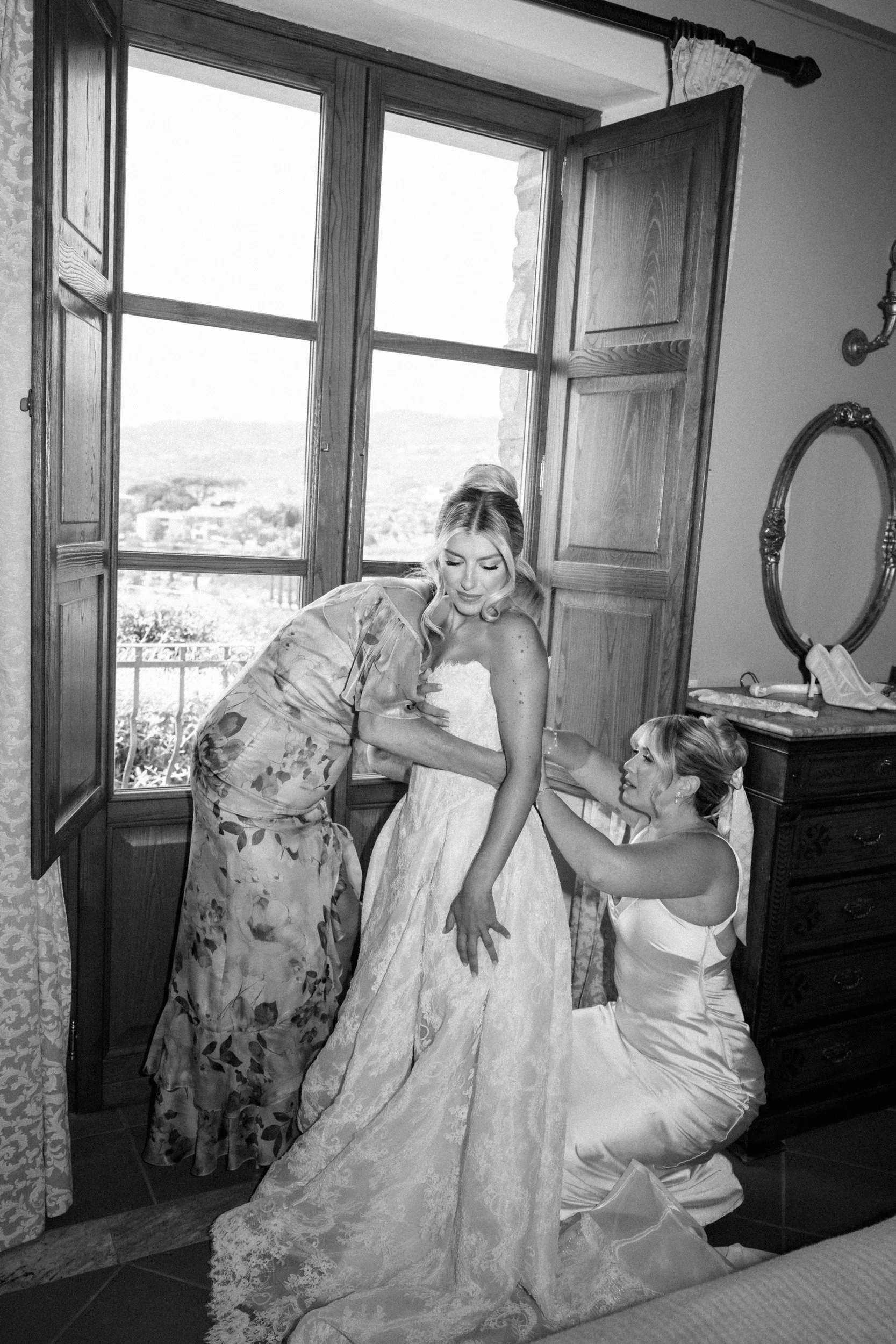 A bride getting help with her wedding dress from two women in a room with large window shutters, overlooking a landscape outside.