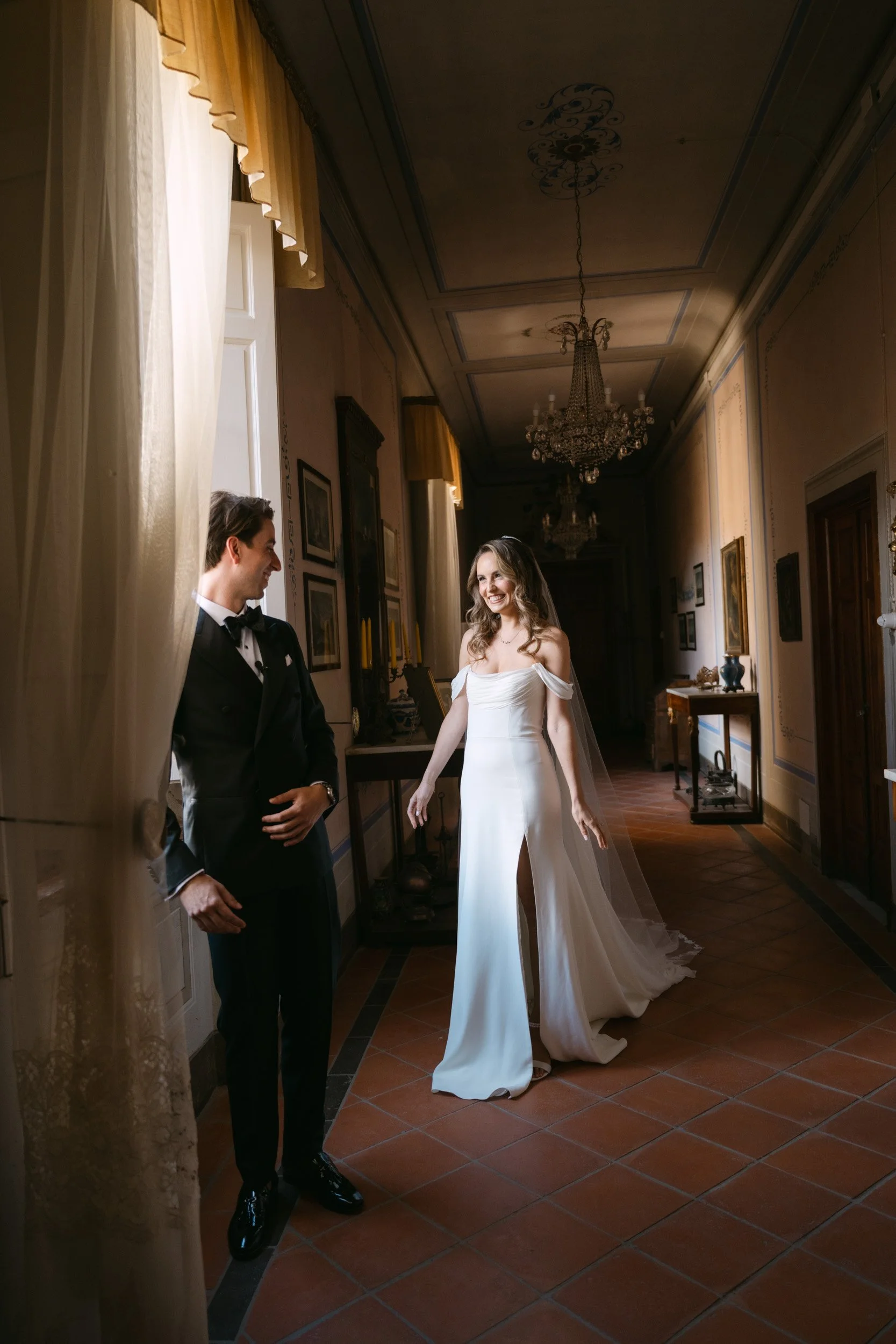 Bride in a white wedding gown with a slit, and groom in a black tuxedo, smiling and looking at each other in an elegant, historic hallway with chandeliers and artwork.