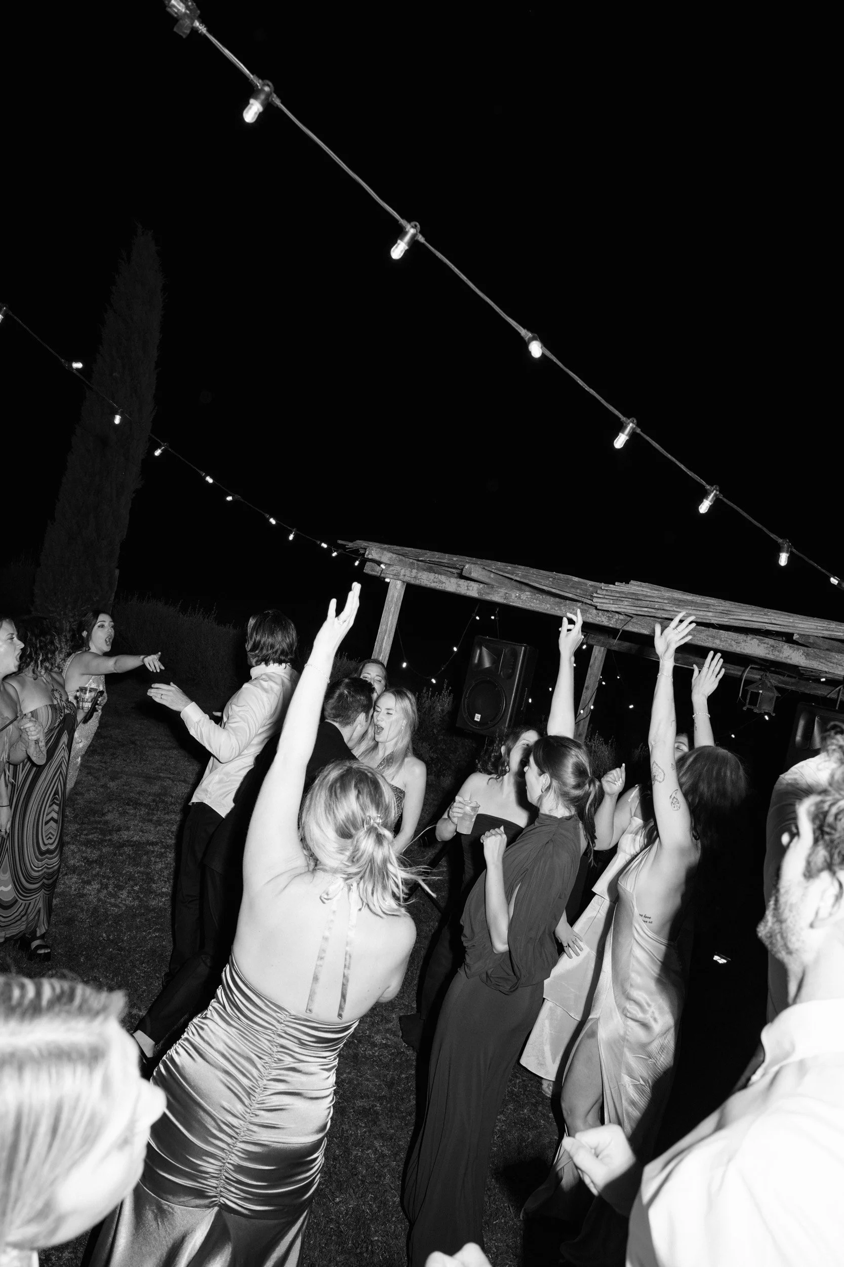 People dancing and celebrating outdoors at night under string lights, with a wooden structure in the background.