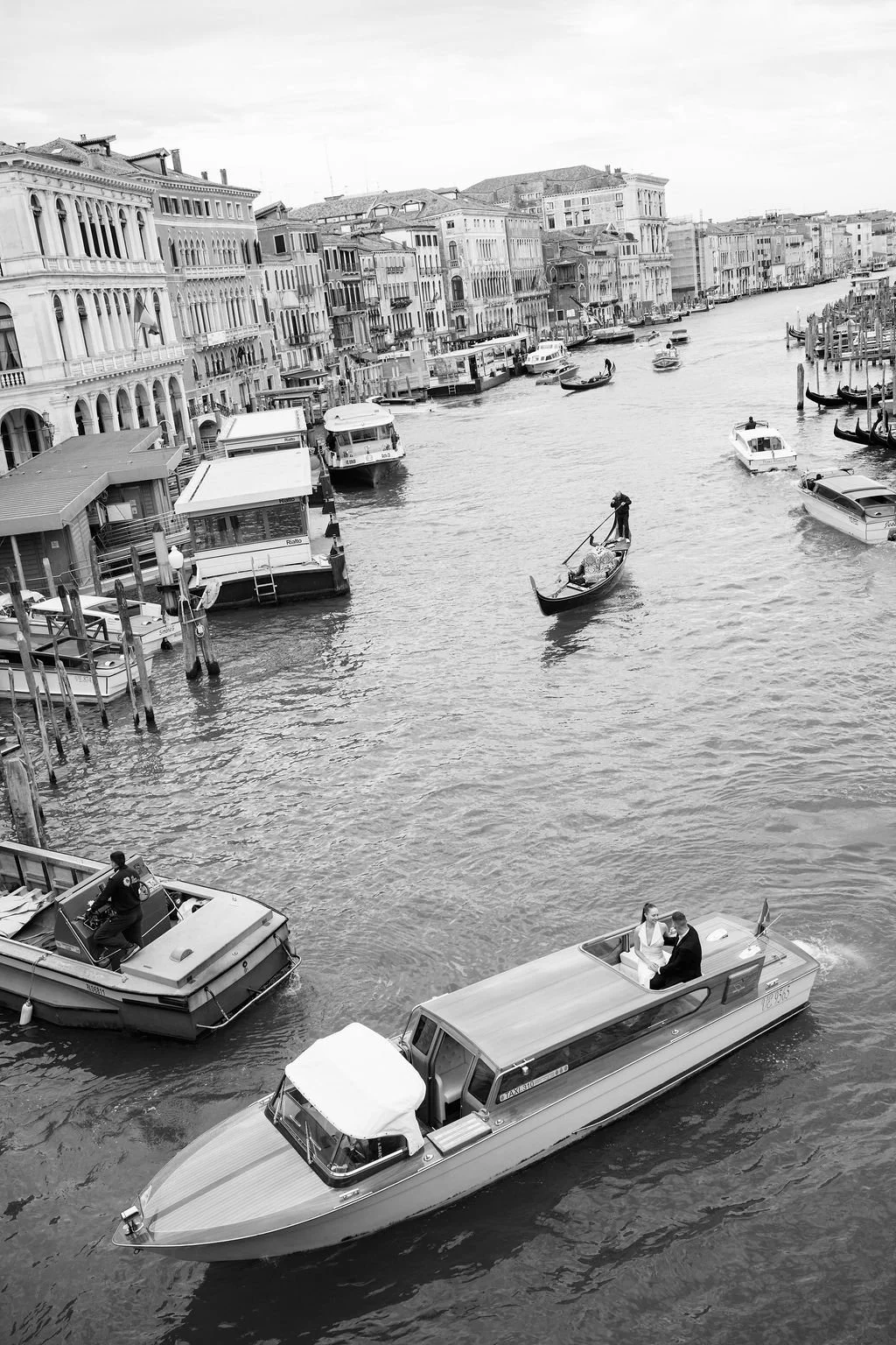 Black and white photo of a canal in Venice with various boats, including a gondola with a gondolier and a motorboat with a couple, building-lined waterway, and cloudy sky.