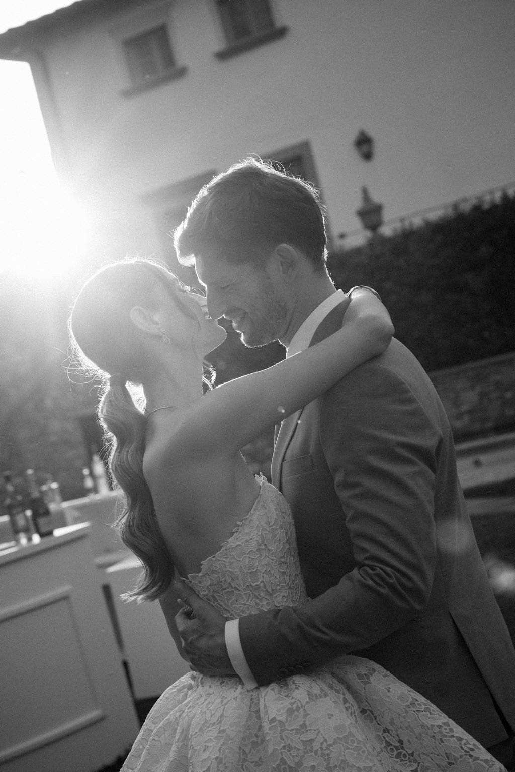 A black and white photo of a couple dancing outdoors, smiling and touching foreheads, with the sun setting in the background.