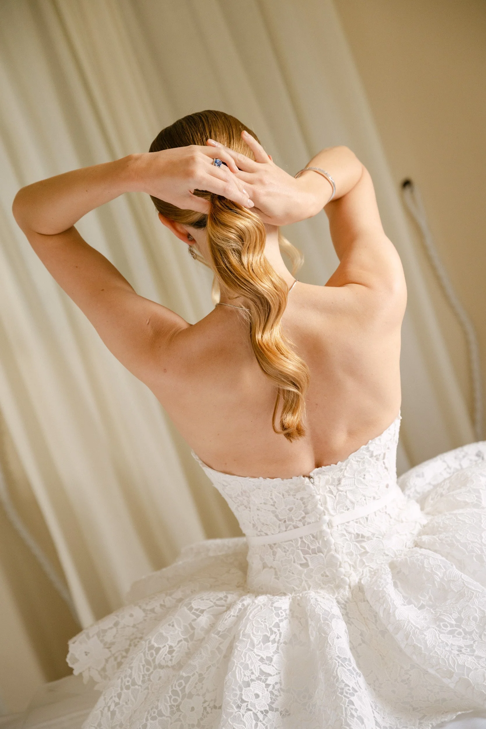 A woman with red hair styled in loose waves, wearing a white lace strapless wedding dress, adjusting her hair with both hands.