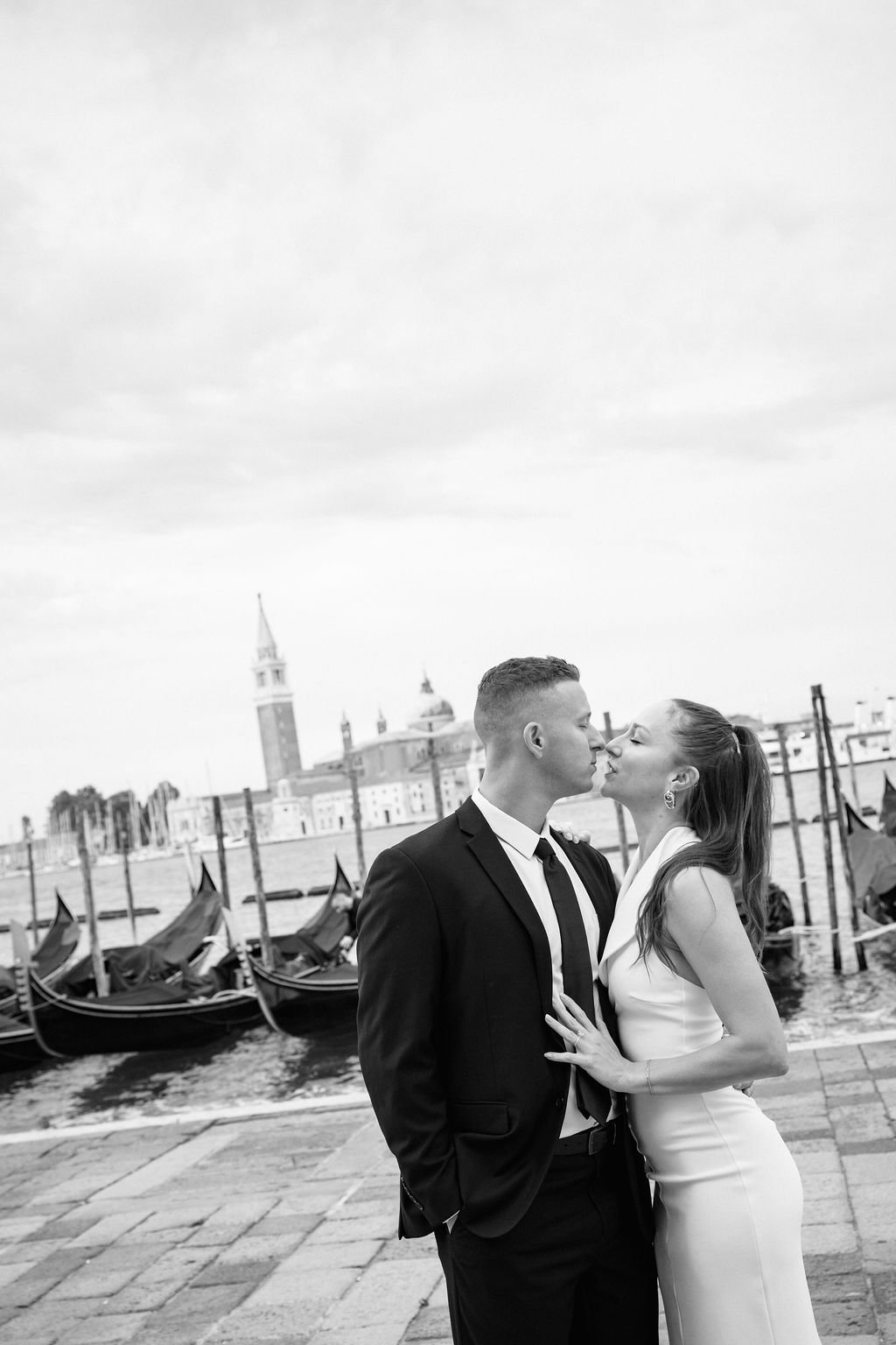 Black and white photo of a couple in wedding attire standing close near a canal with gondolas, with buildings and a church tower in the background, likely in Venice.