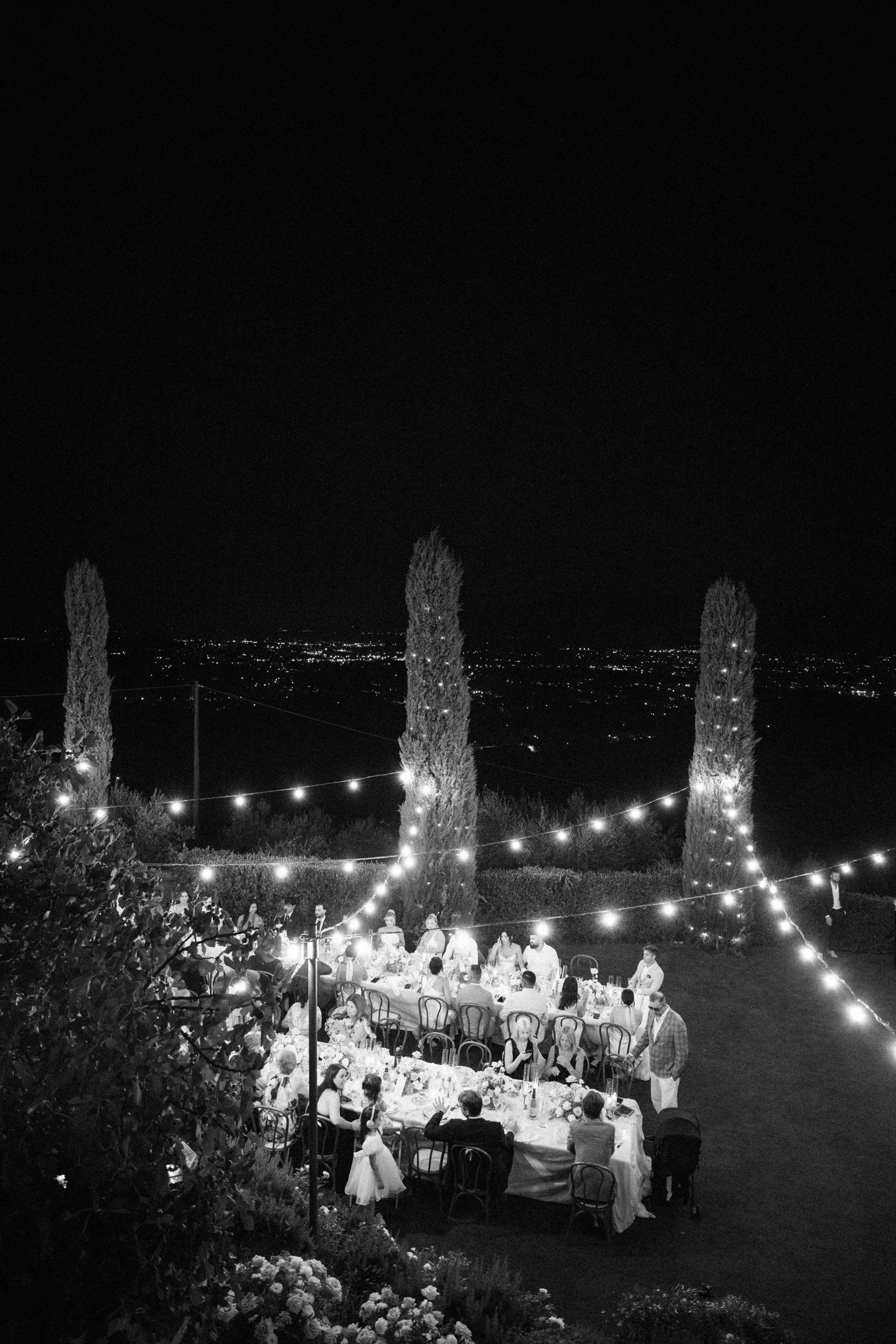 Nighttime outdoor dinner party with people seated at long tables, string lights hanging, tall trees in the background, and city lights in the distance.