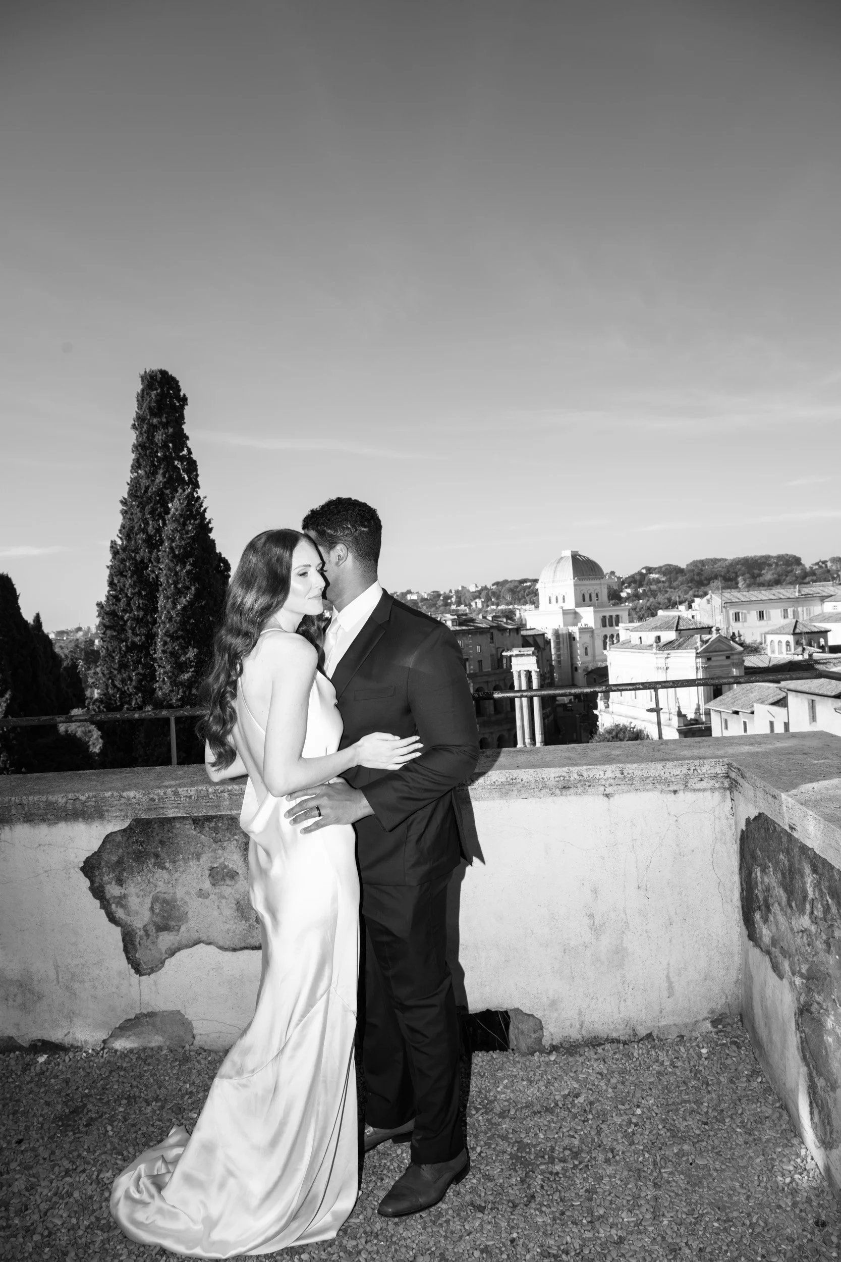 A couple dressed in formal attire, embracing on a rooftop with a cityscape background in black and white.