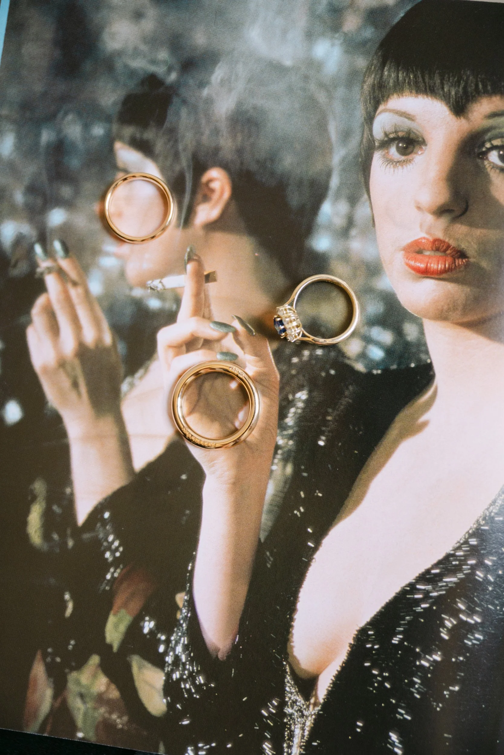 A woman with dark hair and makeup holds a cigarette, with rings resting on a reflective glass surface in front of her. The background is dark and shadowy.