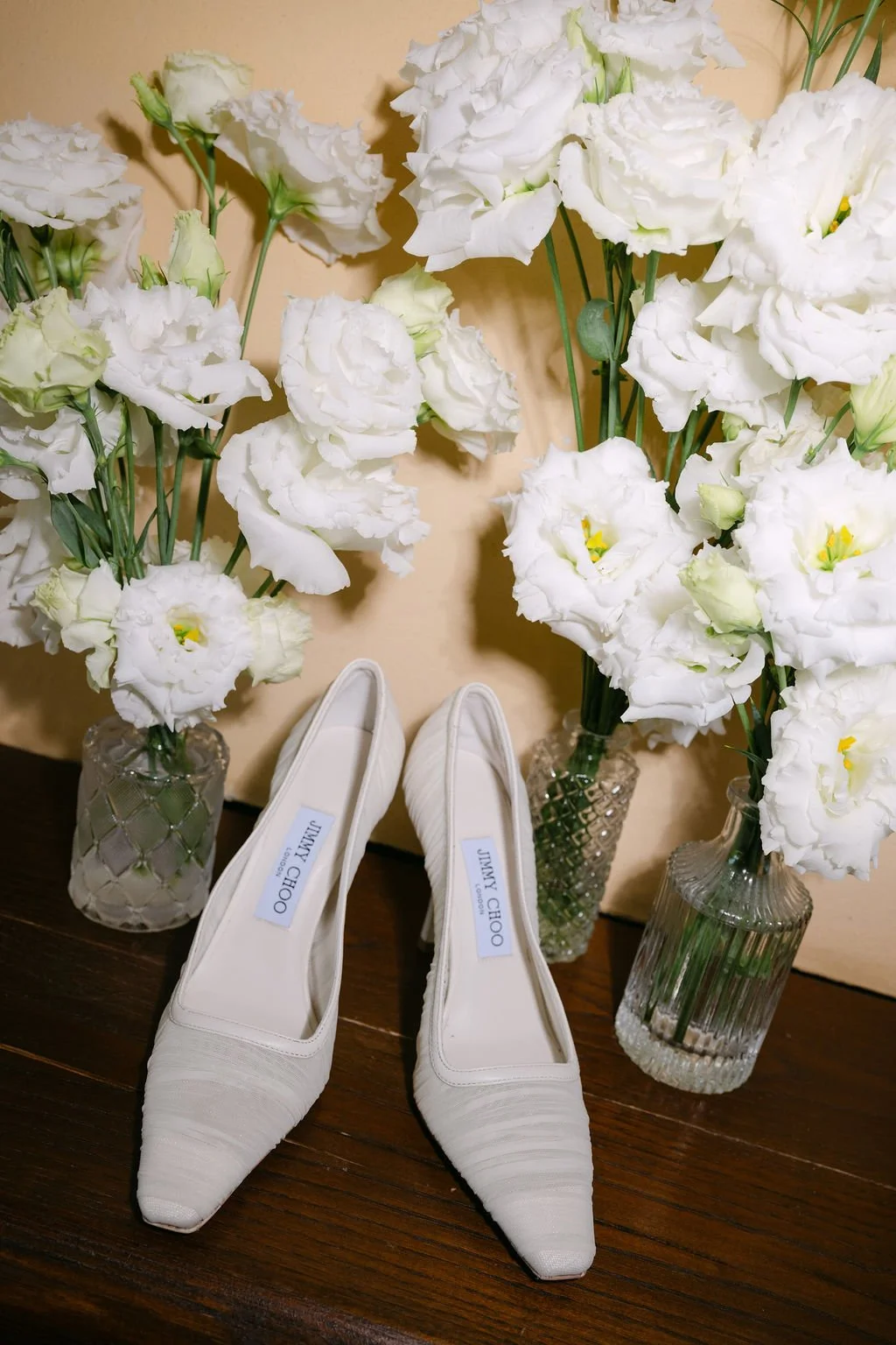 Pair of white Jimmy Choo high heels placed on a wooden surface with three glass vases of white flowers in the background.