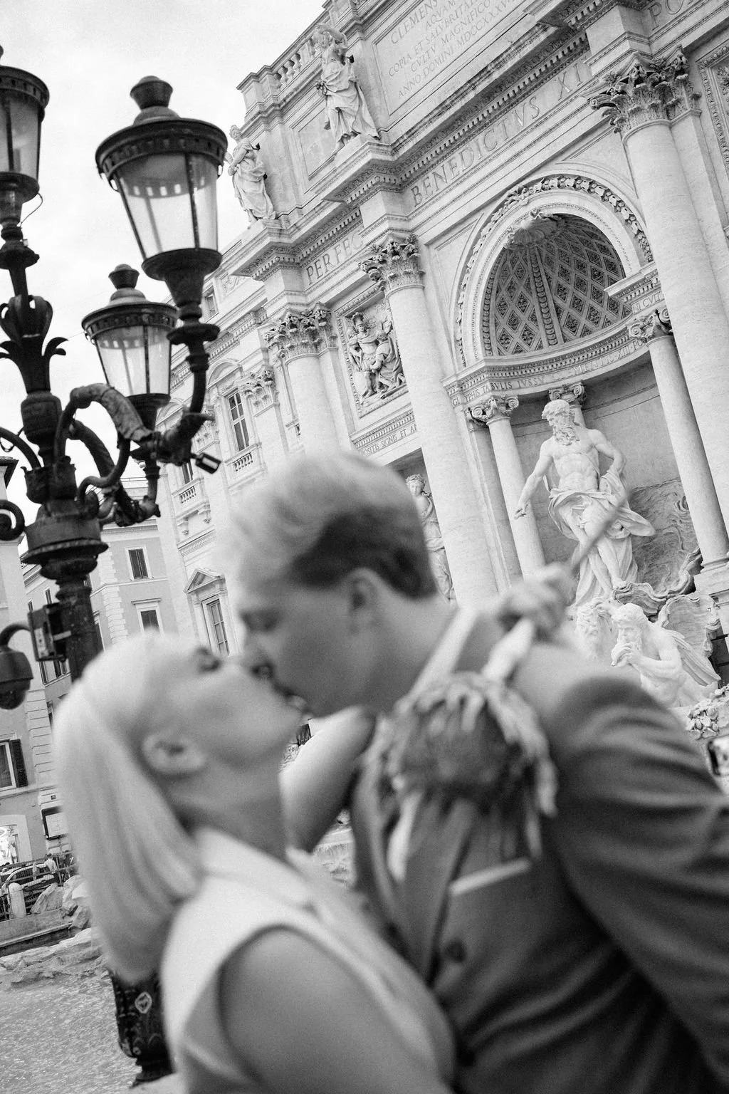A black and white photo of a couple kissing in front of a historic building with classical statues and ornate architectural details, and a vintage street lamp in the foreground.