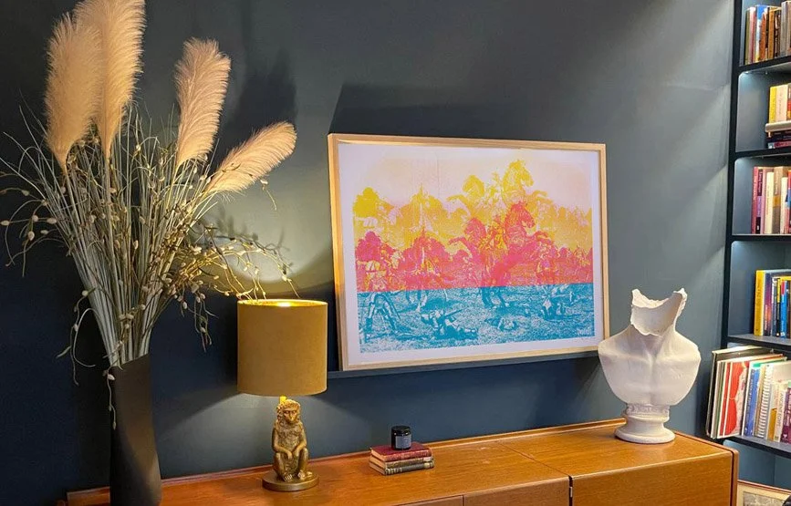 Interior living room with a dark blue wall, a framed colorful print, a tall black vase with pampas grass, a small table lamp with a lion figure base, a stacked set of small books, and a white classical bust sculpture on a wooden sideboard, with a bookshelf on the right.