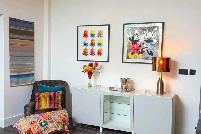 A cozy living room corner features a dark upholstered armchair with colorful cushions, a white sideboard with a glass-door section, a vase of bright flowers, a metallic table lamp, and framed art posters of colorful abstract flowers and cartoon characters on the wall.