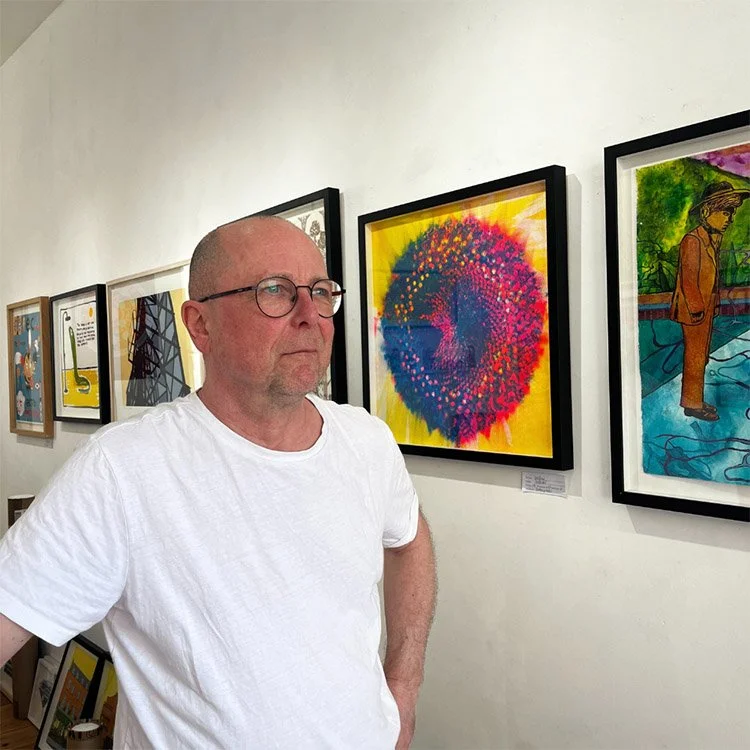A man with glasses and a white t-shirt standing in front of colorful abstract artwork in an art gallery.