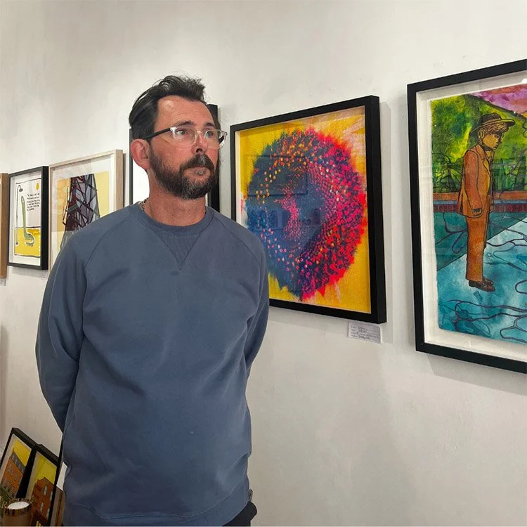 Man with glasses and beard wearing a gray sweatshirt observing colorful abstract art in a gallery.