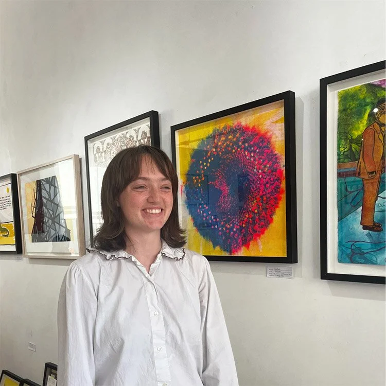 A woman with shoulder-length brown hair and a white shirt smiling in front of colorful abstract artwork at an art gallery.