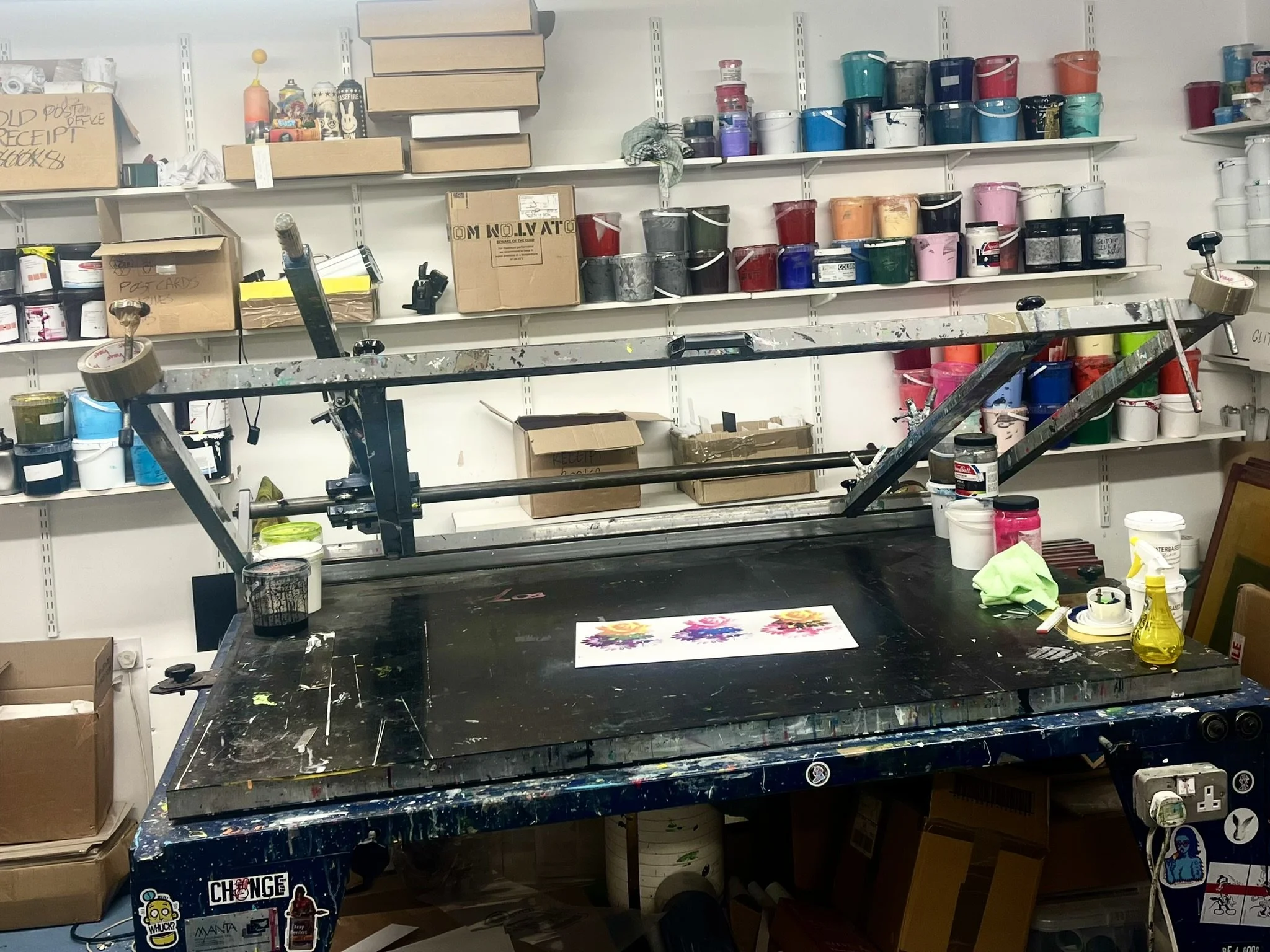 An art studio workspace with a black multi-armed screen printing press on a black table, surrounded by paint containers, brushes, and supplies, with shelves holding colorful paint buckets and cardboard boxes in the background.