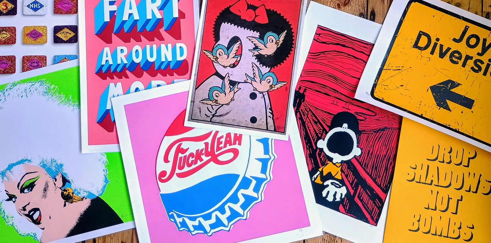 An assortment of colorful art prints and posters with various styles and themes, including cartoon characters, text-based designs, and graphic illustrations lay spread out on a wooden surface.