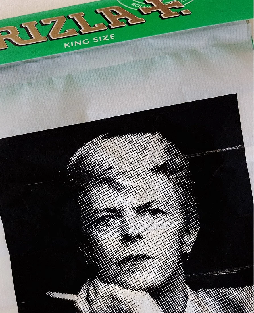 Close-up of artwork by Richard Pendry in a black box frame. A black-and-white halftone portrait of David Bowie smoking a cigarette is screenprinted onto a sheet of Rizla-style rolling papers