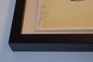 Close-up of a framed picture showing part of a signature and a handwritten note.