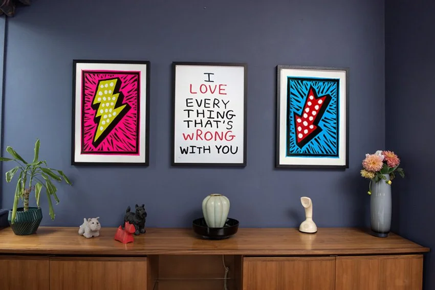 Decorative wall art with three framed pictures, the left with a pink background and yellow lightning bolt, the middle with a quote saying 'I love everything that's wrong with you,' and the right with a blue background and red downward arrow. Below are two small dog figurines, a green potted plant on the left, a white vase in the middle, and a tall gray vase with pink flowers on the right, all placed on a wooden sideboard against a blue wall.