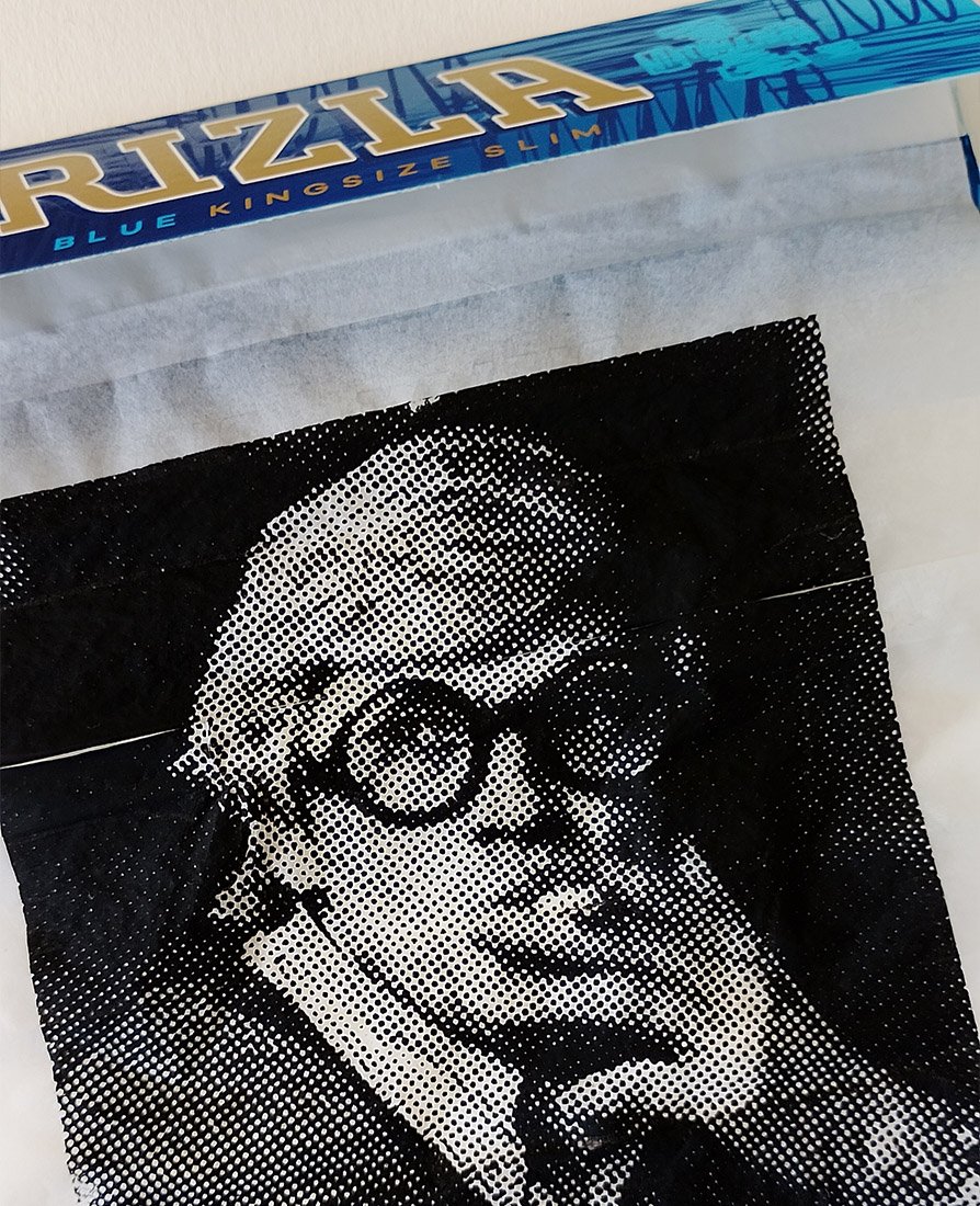 Close-up of artwork by Richard Pendry in a black box frame. A black-and-white halftone portrait of David Hockney smoking a cigarette is screenprinted onto a sheet of Rizla-style rolling papers