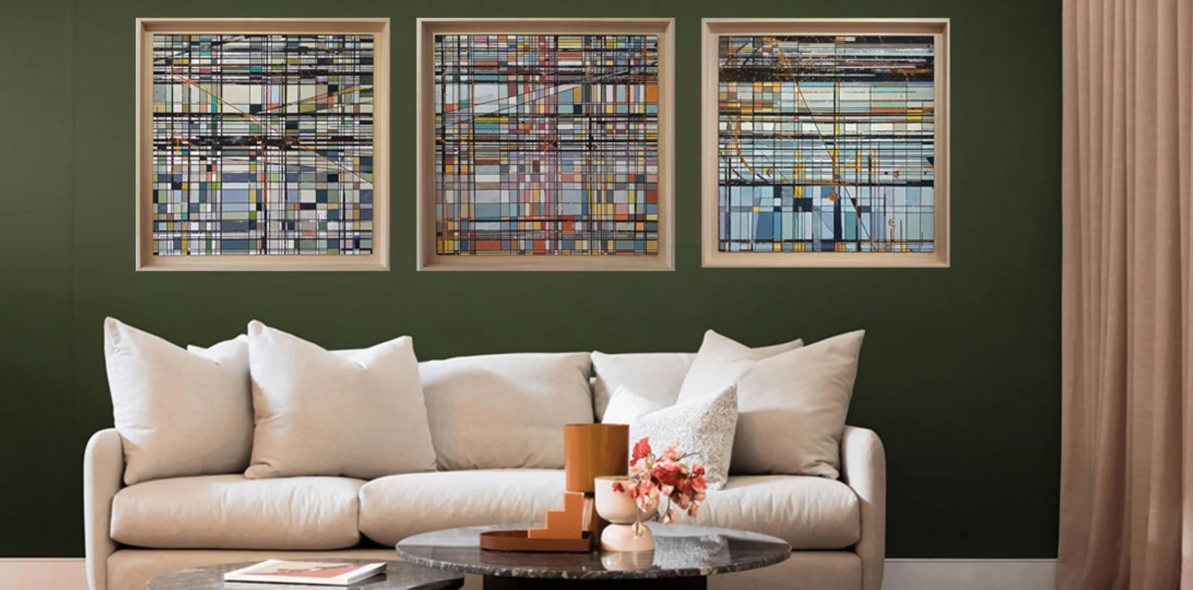 Living room with a green wall, three abstract colorful paintings hanging above a white sofa with multiple pillows, a round coffee table with decorative objects including a vase with pink flowers and a cushion, beige curtains on the right side.