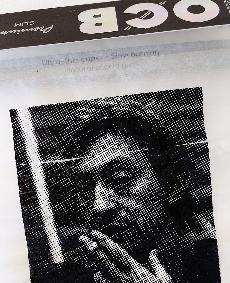 Close-up of artwork by Richard Pendry in a black box frame. A black-and-white halftone portrait of Serge Gainsbourg smoking a cigarette is screenprinted onto a sheet of Rizla-style rolling papers.