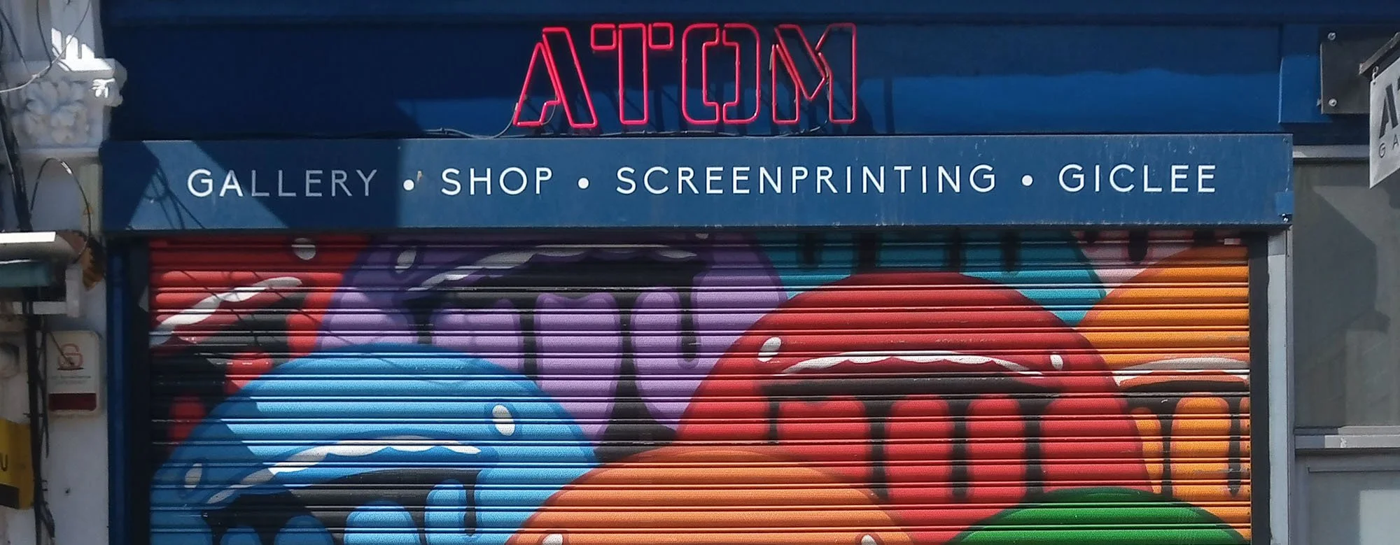 Closed storefront with colorful graffiti artwork, blue canopy with white text offering gallery, shop, screenprinting, and Giclee services, and neon sign reading 'ATCOM' in red.