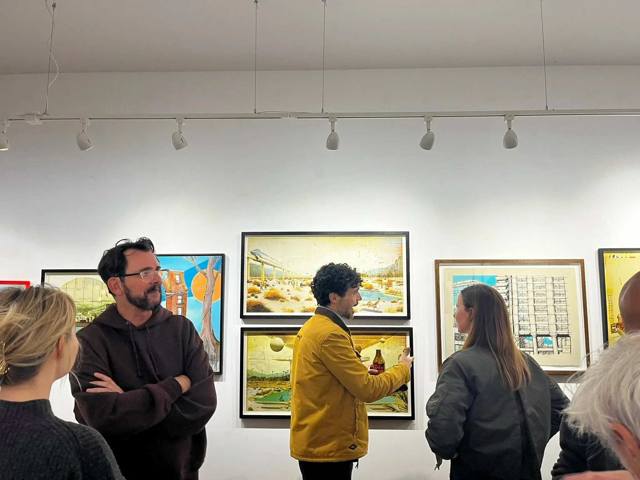 People inside an art gallery looking at framed artwork on a white wall, with one person talking and others listening.