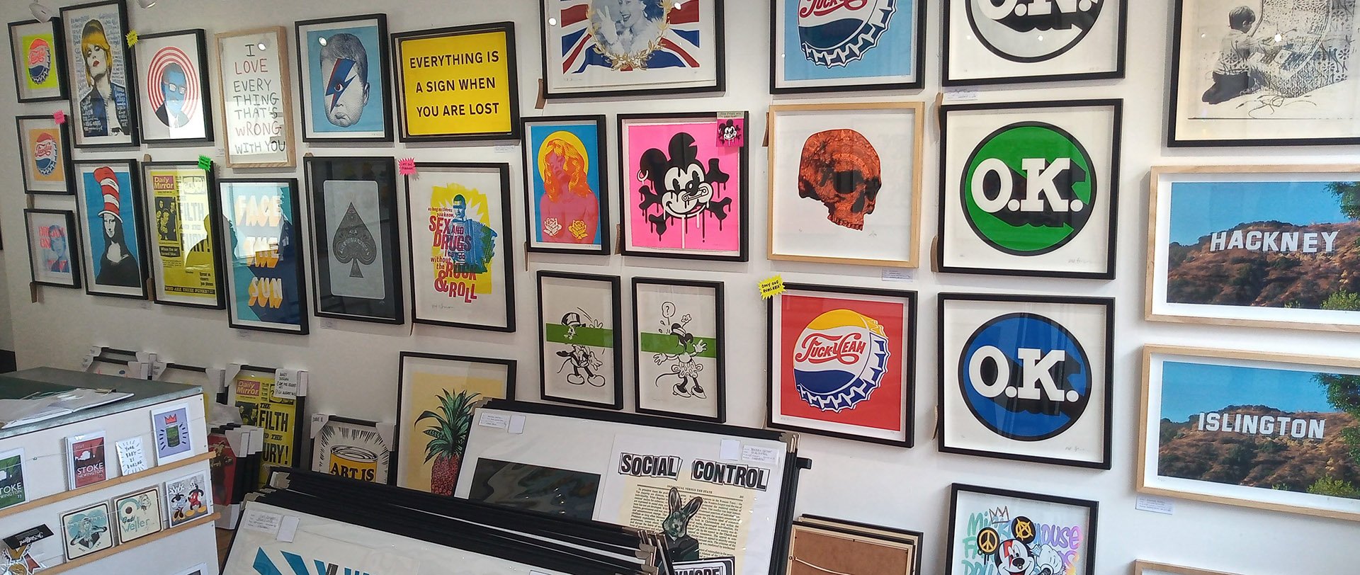 Art gallery or store wall with framed pop art, logos, and images, including O.K. and Hackney. Shelves with prints and signs, some with yellow labels.